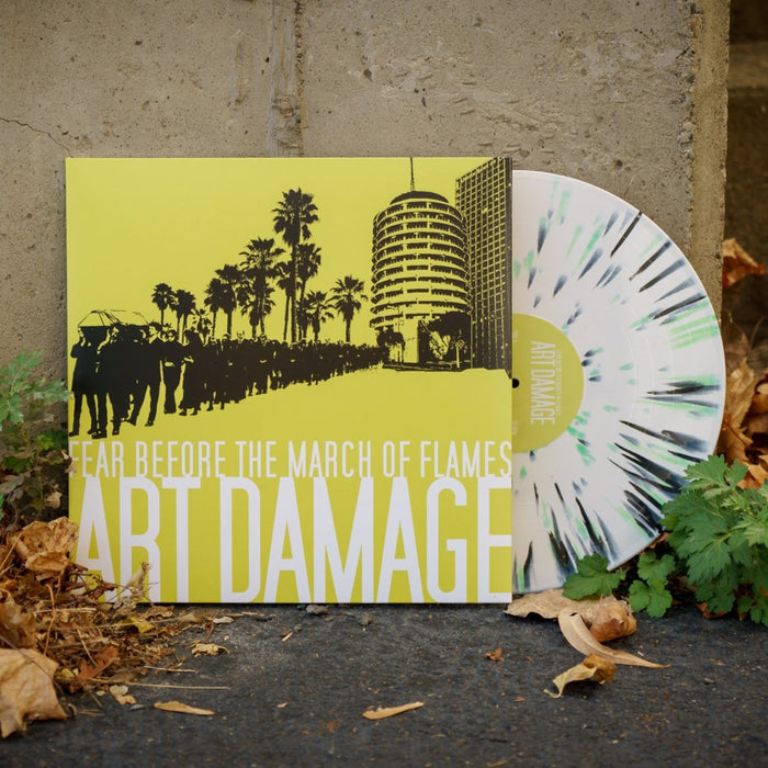 A limited edition LP vinyl with a white and green splatter design from Fear Before is partially pulled from a yellow Art Damage album cover, leaning against a concrete wall among dried leaves and green plants.