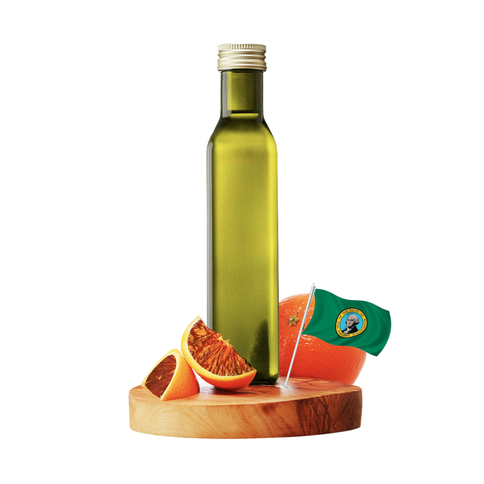 A tall green glass bottle of olive oil with a silver cap, standing on a round wooden board with sliced oranges and a small flag, against a plain white background.