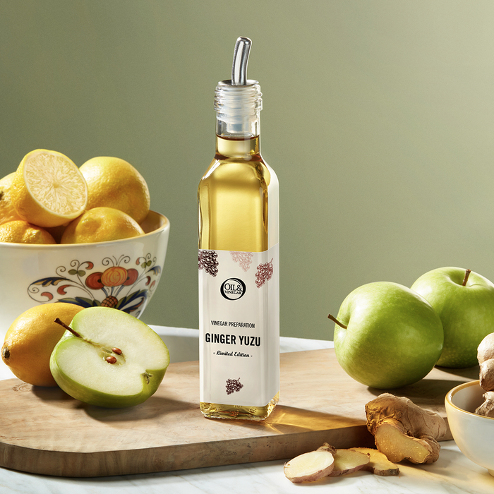 Ginger Yuzu vinegar with a silver topper on a wooden cutting board beside ginger, green apples, and a bowl of lemons