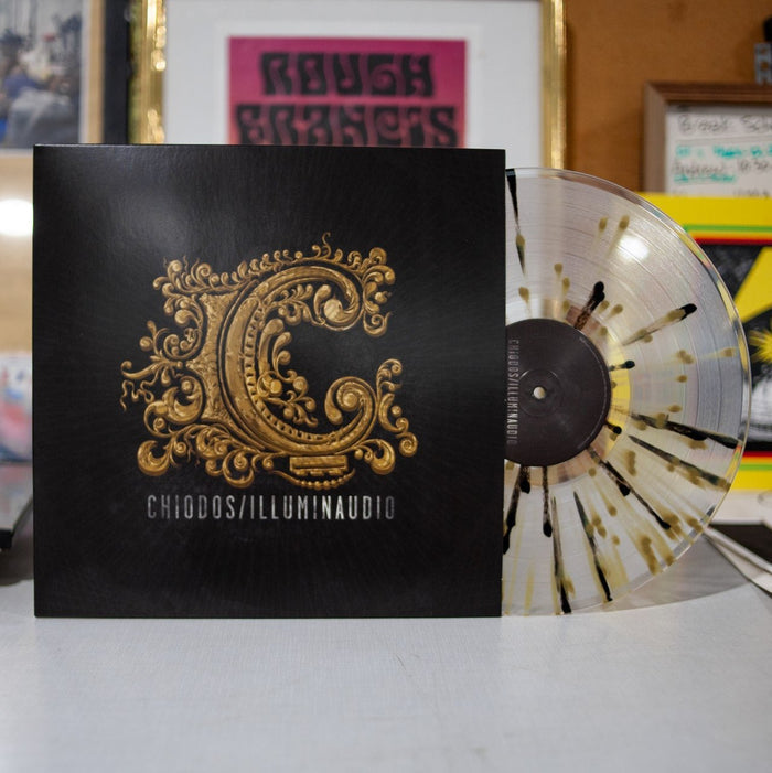 A limited edition Chiodos Illuminaudio vinyl with black and gold ornate C artwork rests on a white surface, showing the clear with gold and black splatter vinyl partially out of its sleeve.