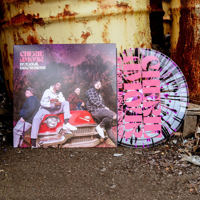 Vinyl jacket with an image of band members sitting on top of a red car. In the background are trees and the night sky, which is tinted pink along with the rest of the cover. In the top left corner there is pink text that says CHERIE AMOUR. Below that is white text that says INTERNAL DISCUSSIONS. Peeking out of the jacket is a clear vinyl with purple and black splatter. Both are on top of a rocky surface and leaning against a rusted surface.