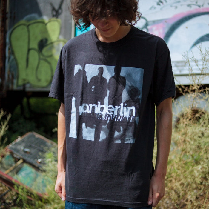 A person with wavy brown hair stands outside wearing the AnberlinInvert T-Shirt by Anberlin, which features a group image and Anberlin text. Blurred graffiti and plants can be seen in the background.
