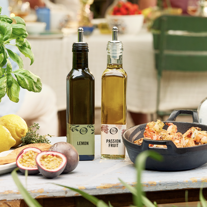 Lemon Olive oil and Passionfruit vinegar on a table outside with greenery, passion fruit slices, whole lemons, and a cast iron dish with cooked shrimp beside it