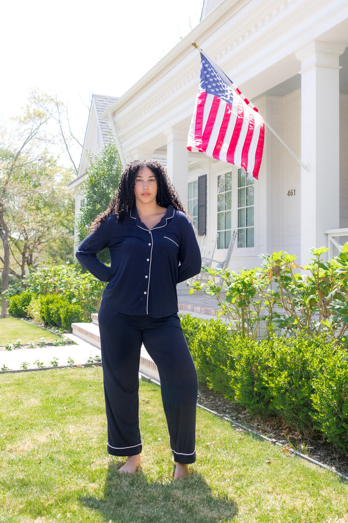 Female wearing the Long-Sleeved Women's Pajama Set in Navy with Cloud Trim standing in the grass in front of a white house wearing the Long-Sleeved Women's Pajama Set in Navy with Cloud Trim