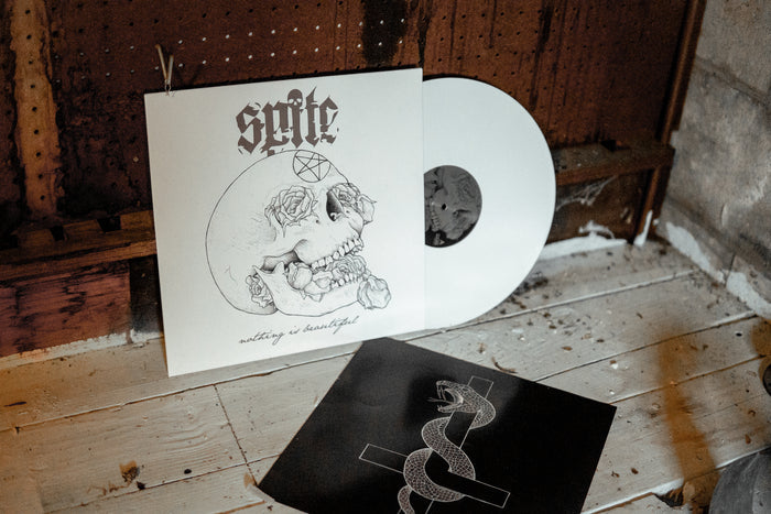 A white jacket shows a skull with roses coming out of its mouth and eyes with the words "nothing is beautiful" at the bottom. "Spite" with a skull as the dot of the "i" is at the top. A white vinyl peeks out of the jacket. The jacket and vinyl rests against a broken down door and floorboard.