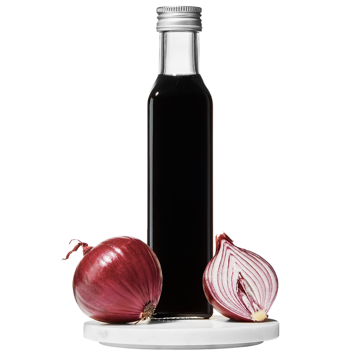 A clear glass bottle of Red Onion Balsamic Vinegar with a cap on top. A whole red onion and a slice of red onion are placed beside the bottle.