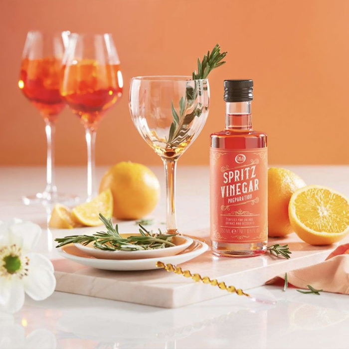 bottle of spritz vinegar next to a wine glass with rosemary on a table with plates and oranges