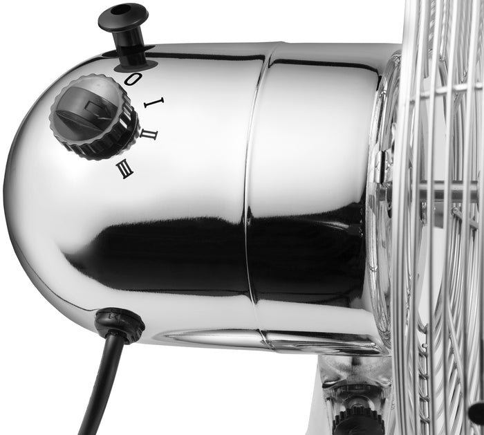 Nahaufnahme des Unold TISCHVENTILATOR CHROME. Hervorzuheben sind die metallische Oberflรคche, der Leistungs- und Geschwindigkeitsregler (Einstellungen 0, I, II, III), das schwarze Netzkabel und ein Abschnitt des Drahtgitters รผber den Ventilatorflรผgeln.