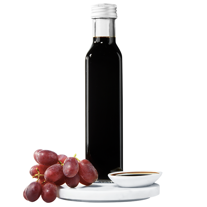 A dark bottle of balsamic vinegar with a silver cap, accompanied by a small bunch of red grapes.