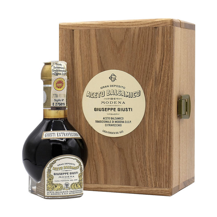 A 100 ml bottle of Aceto Balsamico Tradizionale di Modena DOP vinegar, placed in front of its packaging box. The box is made of wood and has a label displaying the brand and product name.
