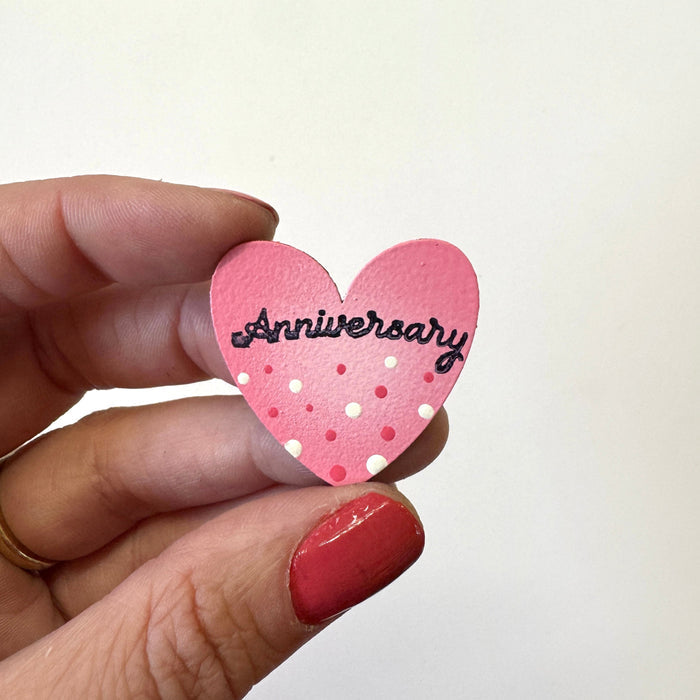 A hand holds a small pink heart-shaped magnet that reads 'Anniversary' in black script, decorated with white and pink polka dots, against a plain off-white background.