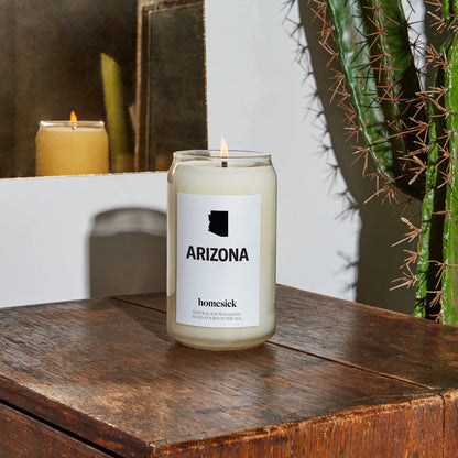 A close up of a lit Arizona candle on a wooden table. In the corner, one can see a cactus.