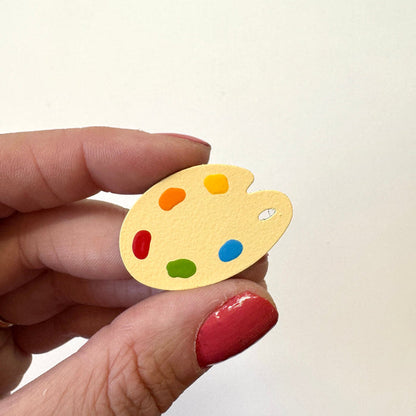 A hand holds a small yellow artist palette magnet with colorful paint dots against a plain off-white background.