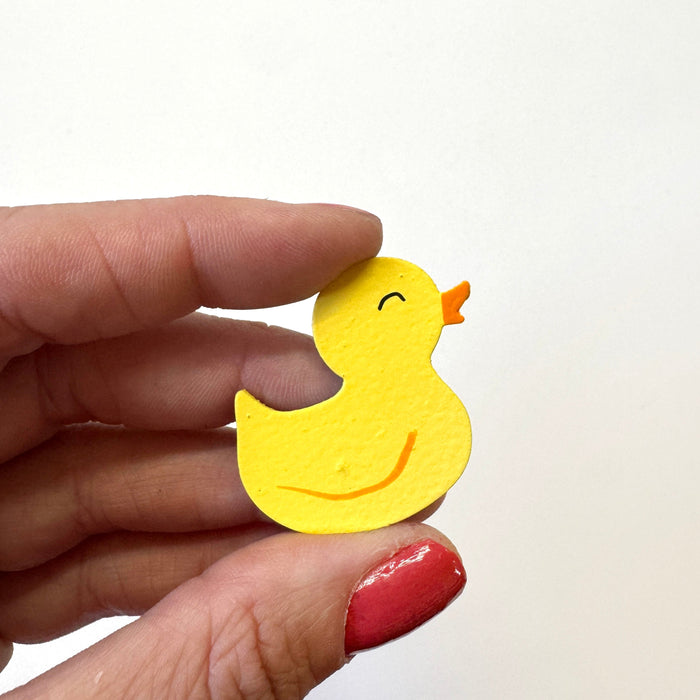 A small yellow rubber duck magnet held between a person's fingers against a plain white background.