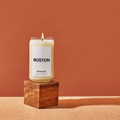A Boston Homesick Candle displayed on top of a wooden cube. The cube is displayed on a cork surface and reddish background.