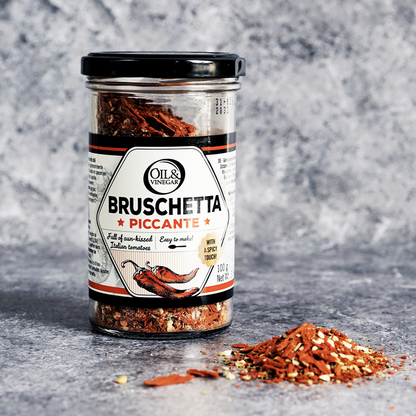 Transparent jar of bruschetta piccante with a black, white and red label with peppers on it indicating the flavor is spicy. All on a white background