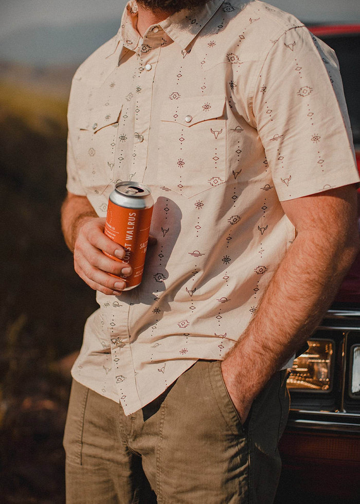 Man in patterned shirt holds a can, standing by a car. Sunlight casts shadows, creating a relaxed, outdoor vibe.