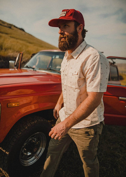 Bull Shirt - Iron & ResinaMan in a red cap and patterned shirt stands by a vintage red truck in a grassy field, looking into the distance.