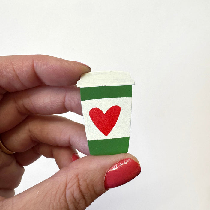 A hand holds a miniature white paper coffee cup magnet featuring green stripes and a red heart, against a plain white background.