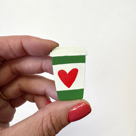 A hand holds a miniature white paper coffee cup magnet featuring green stripes and a red heart, against a plain white background.