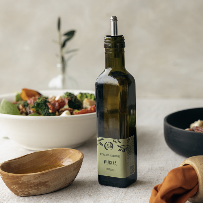 Puglia extra virgin olive oil with a silver topper on a white table next to a olive wood bowl with veggies in the background