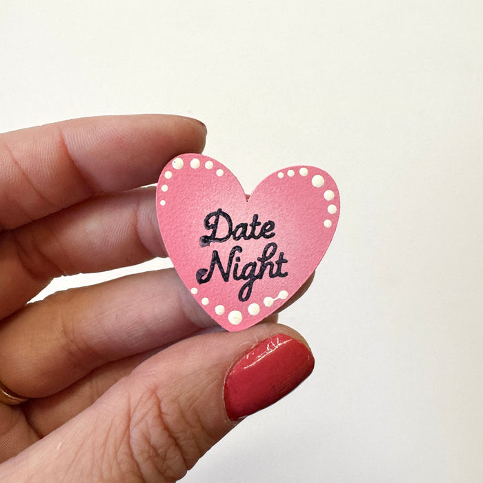 A hand holds a small pink heart-shaped magnet that reads 'Date Night' in black script, against a plain off-white background.