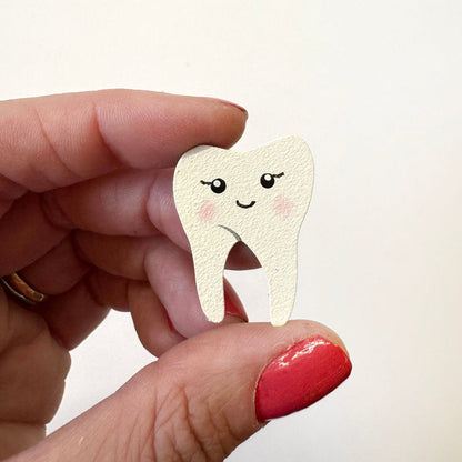A hand with red nail polish holds a small, cute, tooth-shaped magnet with a smiling face against a plain white background.