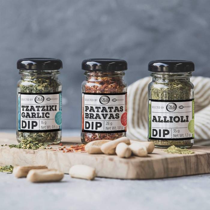 Three small jars of different flavors of dried herbs (Tzatziki garlic, Patatas Bravas, and Allioli)on a wooden board on a gray background