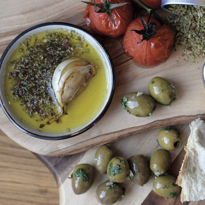 Wooden board with olives, tomatoes, and a dish with olive oil, herbs, and a garlic clove.