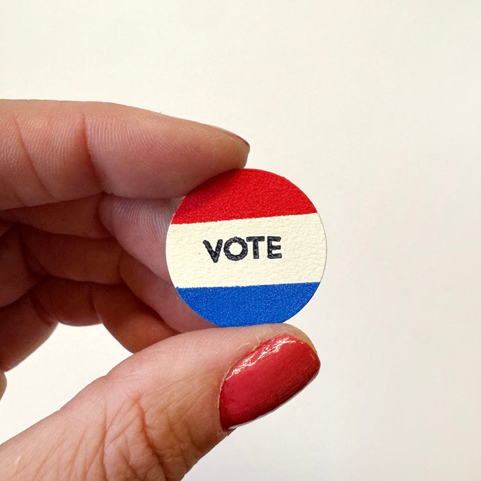 A hand holds a small round magnet featuring red, white, and blue horizontal stripes with the word 'VOTE' printed in black in the center.