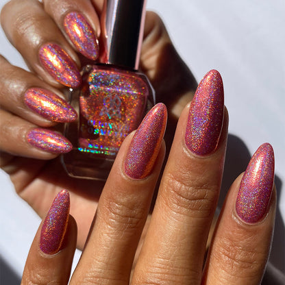 Close-up of nails painted with Flamin' Hot Holo Nail Lacquer on medium skin tone. Hand is holding polish bottle.