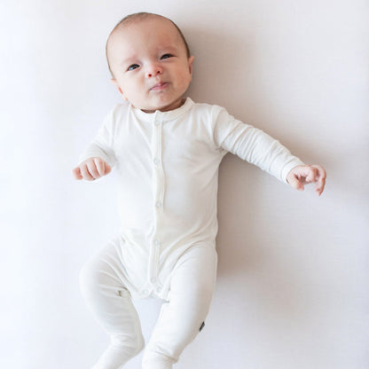 Baby wearing Kyte Baby Snap Footie in Cloud. Laying on white surface and looking at the camera