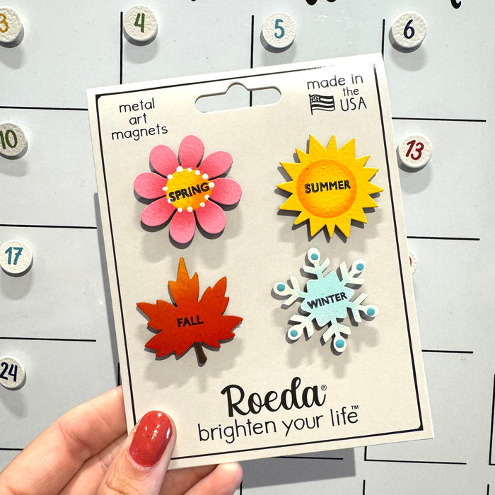 A hand holds a package of Roeda metal art magnets featuring seasonal designs: a pink spring flower, a yellow summer sun, an orange autumn leaf, and a light blue winter snowflake, set against a white magnetic calendar board.