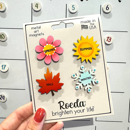 A hand holds a package of Roeda metal art magnets featuring seasonal designs: a pink spring flower, a yellow summer sun, an orange autumn leaf, and a light blue winter snowflake, set against a white magnetic calendar board.