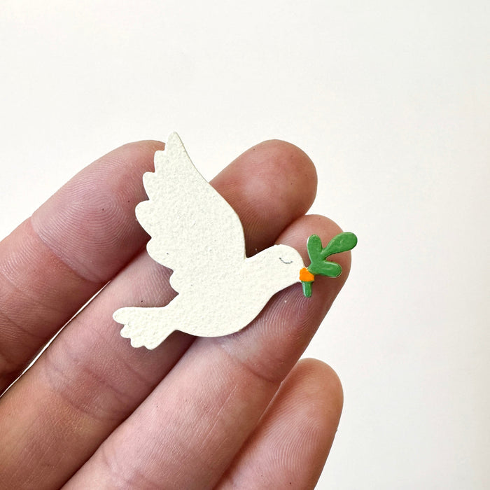 A hand holding a small white dove-shaped magnet with a green leafy sprig and orange detail on its beak, against a plain light background.