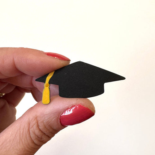 A miniature black graduation cap magnet with a yellow tassel held between fingers  against a plain white background.