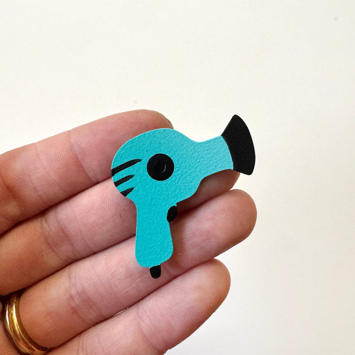 A small turquoise and black hair dryer-shaped magnet held between fingers against a plain off-white background.