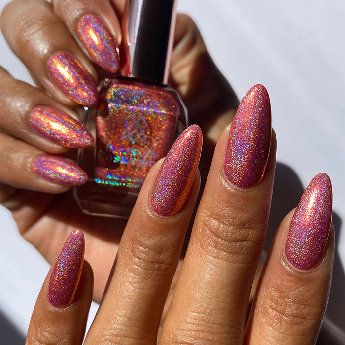 Close-up of nails painted with Flamin' Hot Holo Nail Lacquer on medium skin tone. Hand is holding polish bottle.