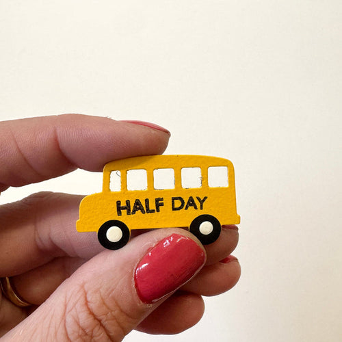 A hand holds a small yellow school bus magnet with the words 'HALF DAY' in black text, against a plain off-white background.