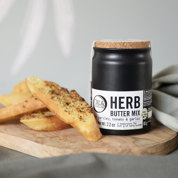 Ceramic jar of herb butter mix with a cork lid on a wooden surface. With toasted herb buttered bread beside it on a gray background.
