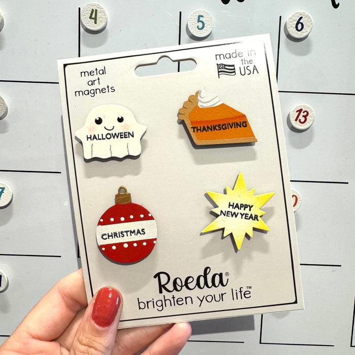 A hand holds a package containing four Roeda metal art magnets with Halloween, Thanksgiving, Christmas, and Happy New Year designs, set against a white magnetic calendar board.