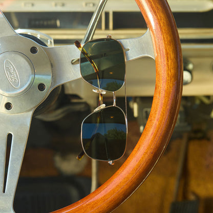 hanging on a steering wheel of a car diff eyewear jonas aviator prescription sunglasses with a gold frame and g15 lenses front view