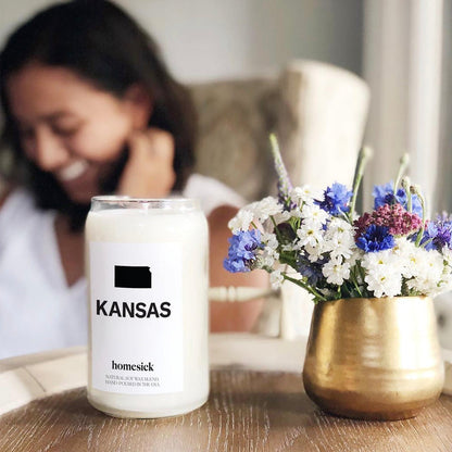 A close up shot of a Kansas Homesick candle on the left and a small flower arrangement in a gold pot on the right. The background is blurry but there is a woman sitting in a chair.