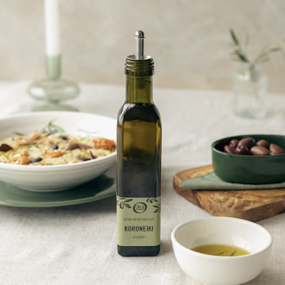 Koroneiki extra virgin olive oil on a white table next to a bow of olives and a small bowl of olive oil