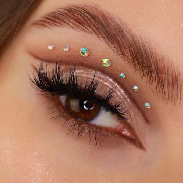 Shimmery nude‑champagne eyelid shows ColourPop Ladybird Super Shock Shadow sparkling across the lid, paired with long lashes, soft winged liner, and iridescent rhinestones along the brow bone in a glam close-up.