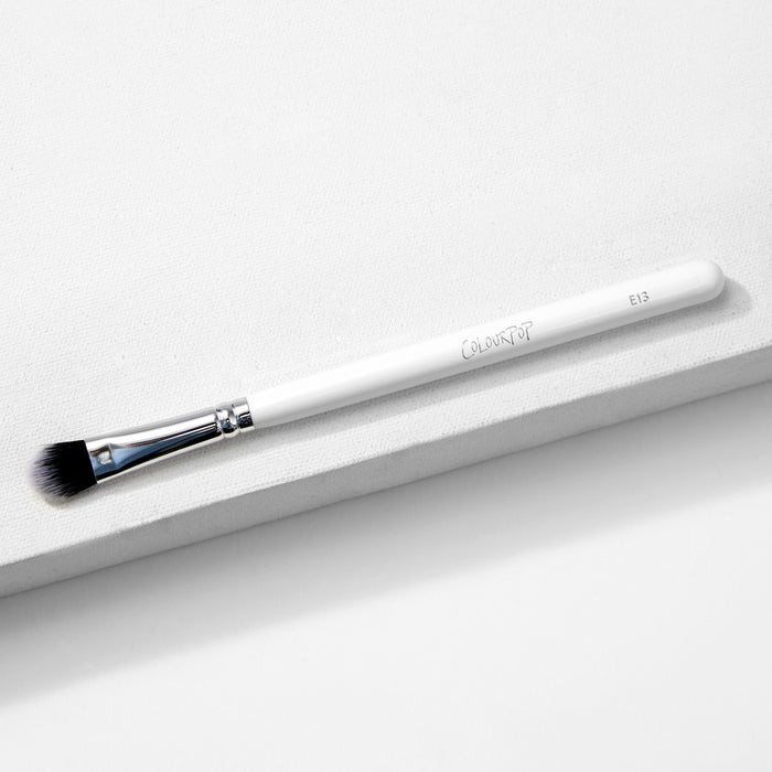 Large Shader Brush makeup brush by ColourPop lies diagonally on a white textured surface, showcasing a white handle with logo and E13, a shiny silver ferrule, and flat dense black-to-white bristles designed for packing and shading eyeshadow.