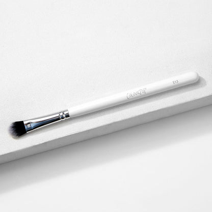 Large Shader Brush makeup brush by ColourPop lies diagonally on a white textured surface, showcasing a white handle with logo and E13, a shiny silver ferrule, and flat dense black-to-white bristles designed for packing and shading eyeshadow.