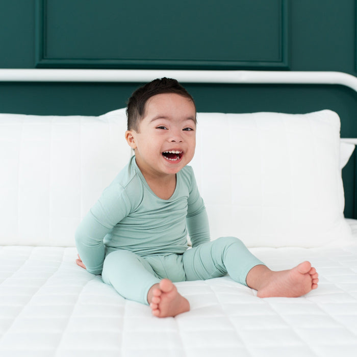 Smiling toddler sitting on a white bed while wearing Long Sleeve Pajama Set in Sage