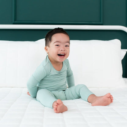 Smiling toddler sitting on a white bed while wearing Long Sleeve Pajama Set in Sage
