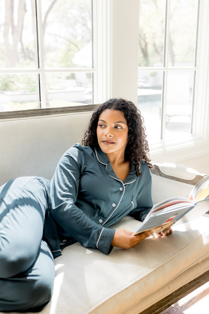 Female sitting on a couch underneath a window with a book in her hand wearing the Long-Sleeved Women's Pajama Set in Atlantic with Cloud Trim